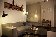 Ein graues Sofa steht neben einem dunklen Couchtisch in einem Wohnzimmer mit warmer Beleuchtung. Im Hintergrund sorgen eine mehrflammige Stehleuchte sowie eine Tischlampe auf einem weißen Sideboard für indirektes Licht.